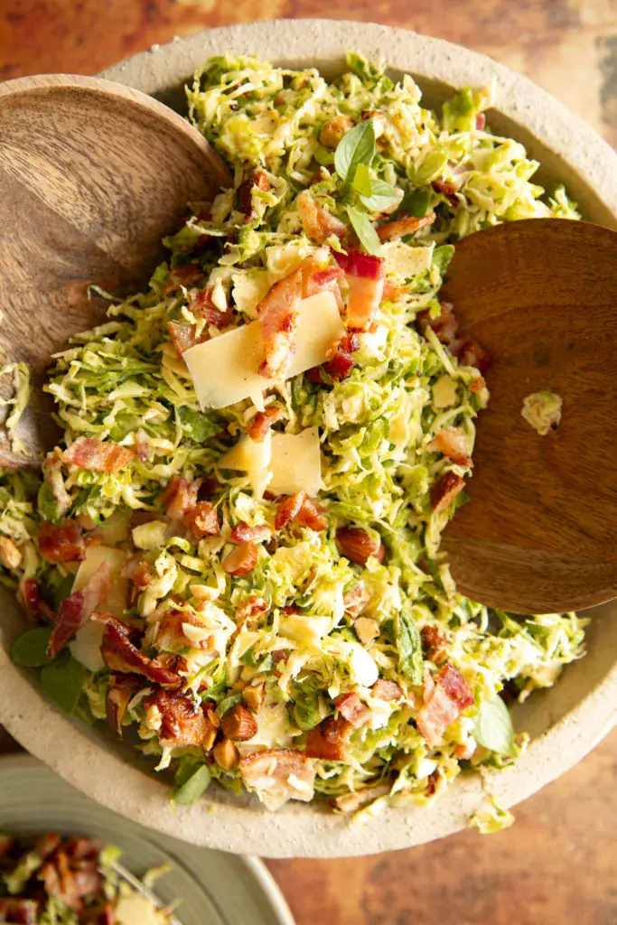 A serving of Brussels sprouts salad plated with bacon, shaved parmesan, almonds, and herbs on a green ceramic dish with a fork.
