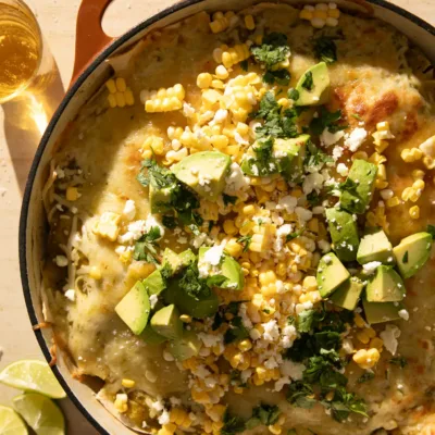 Close-up of a serving spoon scooping out a cheesy portion of green chile enchilada skillet topped with avocado, corn, and cilantro.