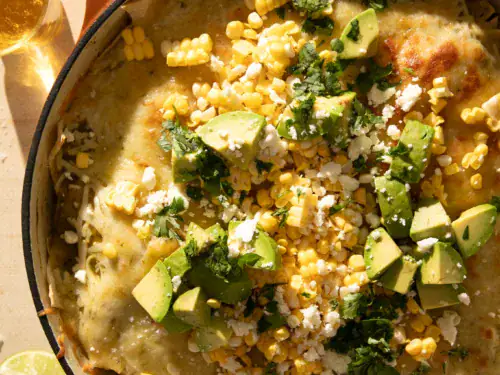 Close-up of a serving spoon scooping out a cheesy portion of green chile enchilada skillet topped with avocado, corn, and cilantro.