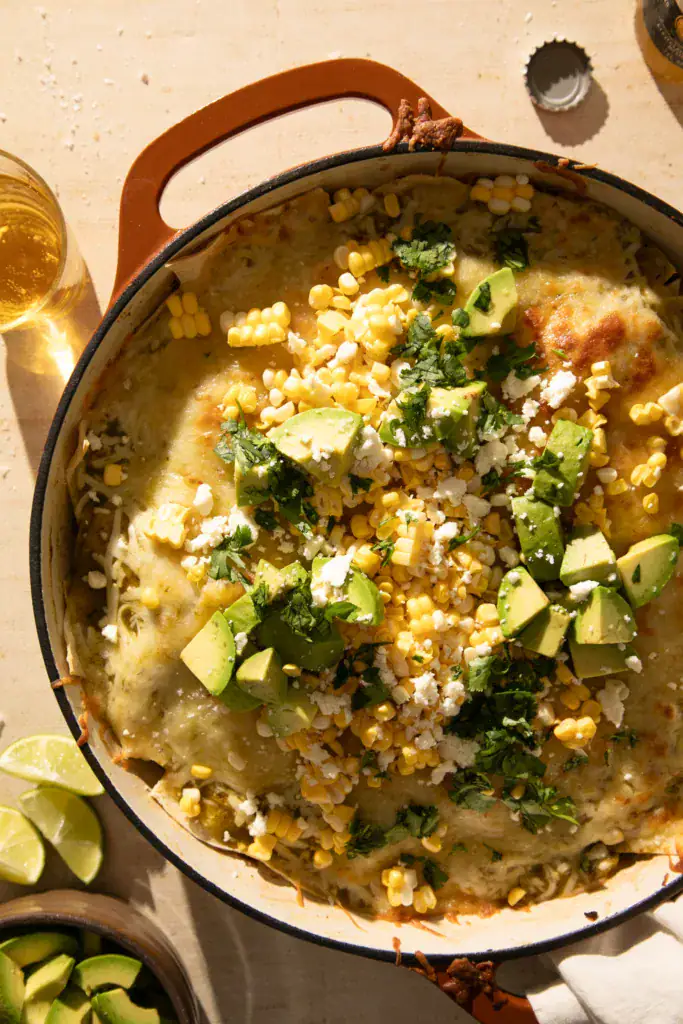 Close-up of a serving spoon scooping out a cheesy portion of green chile enchilada skillet topped with avocado, corn, and cilantro.