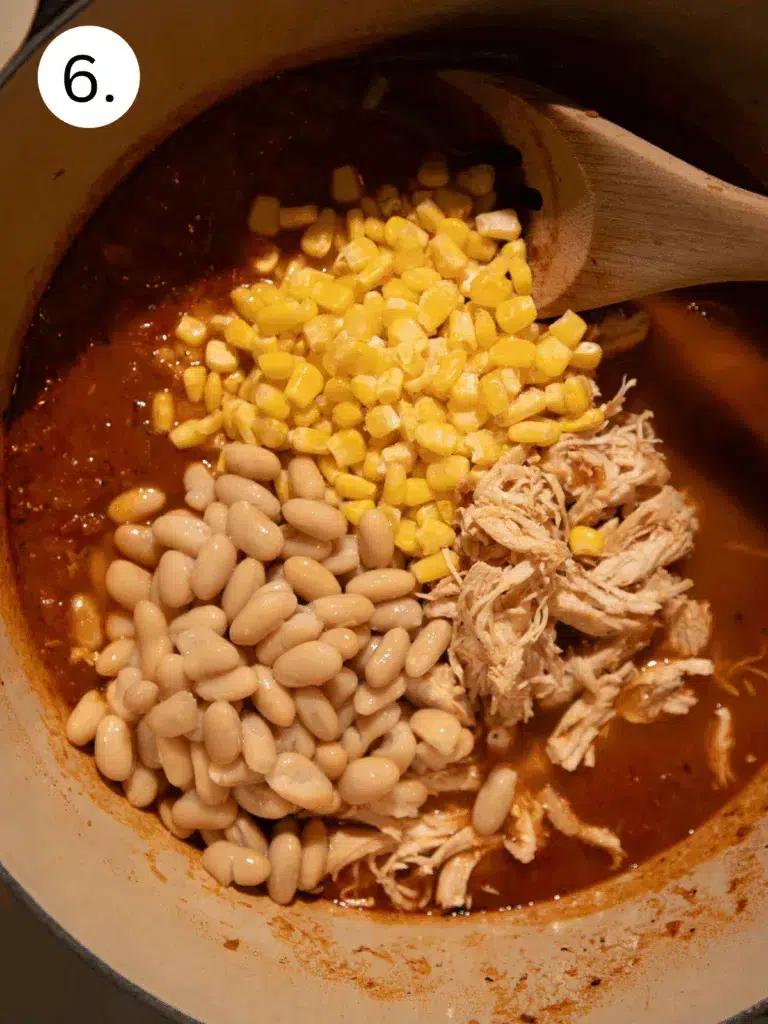 Pot of chili with shredded chicken, sweet corn, and white beans being stirred into the smoky tomato broth.