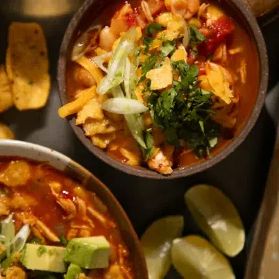 Chili served in a rustic bowl topped with shredded cheese, green onions, cilantro, and corn chips with lime wedges on the side.