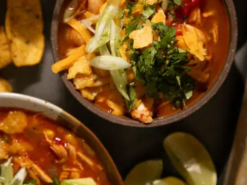 Chili served in a rustic bowl topped with shredded cheese, green onions, cilantro, and corn chips with lime wedges on the side.