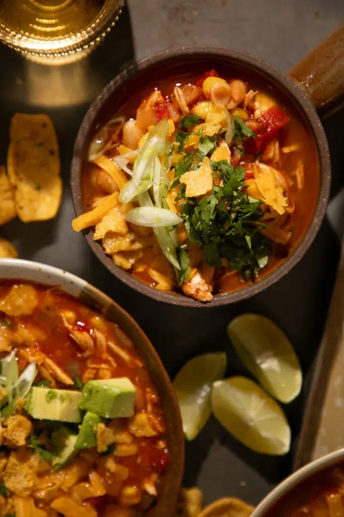 Chili served in a rustic bowl topped with shredded cheese, green onions, cilantro, and corn chips with lime wedges on the side.