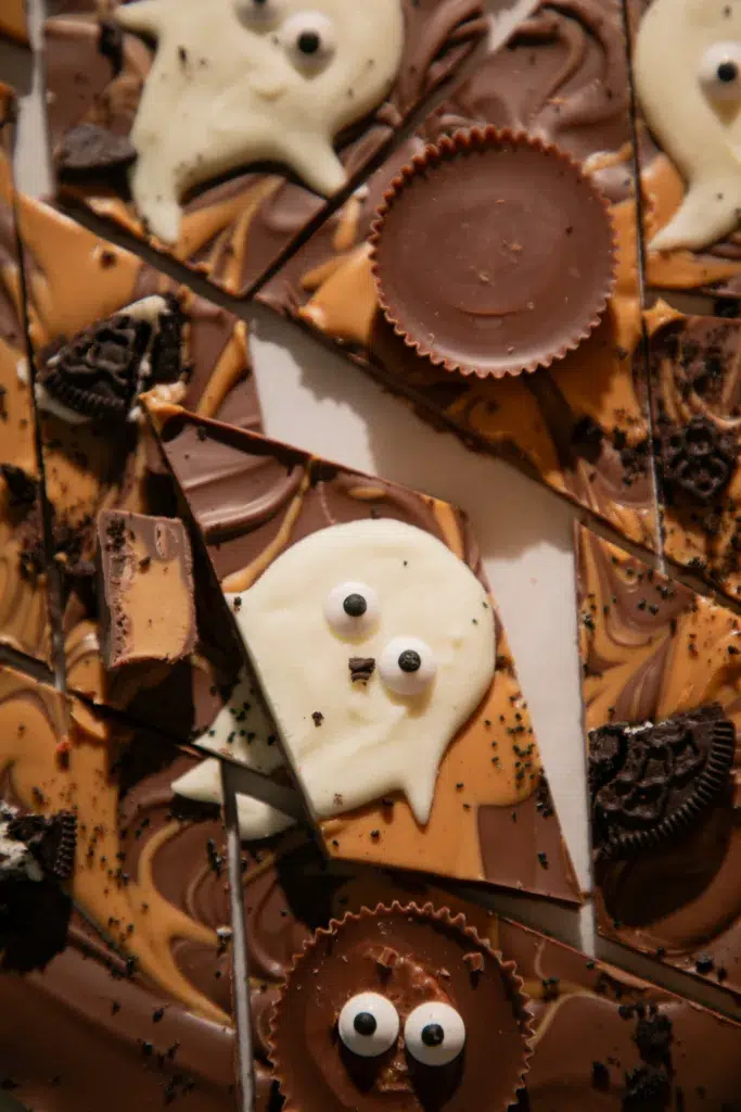 Pieces of Halloween Peanut Butter Chocolate Ghost Bark topped with white chocolate ghosts, Reese’s Cups, and crushed Oreos.