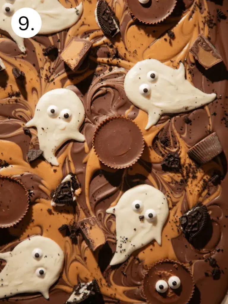 Halloween bark decorated with peanut butter swirls, Reese’s Cups, Oreo pieces, and white chocolate ghosts with candy eyes before chilling.