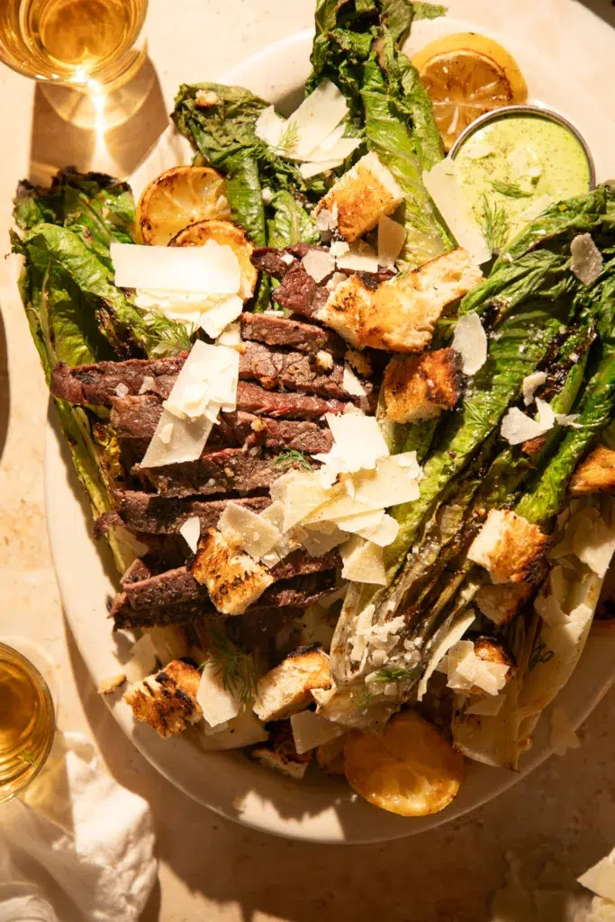 Overhead view of grilled romaine topped with sliced ribeye, parmesan shavings, grilled croutons, and green herb Caesar dressing.