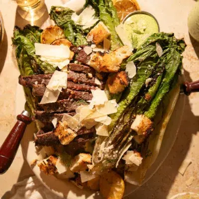 Close-up of charred romaine hearts topped with ribeye steak slices, parmesan, croutons, and a drizzle of creamy herb dressing.