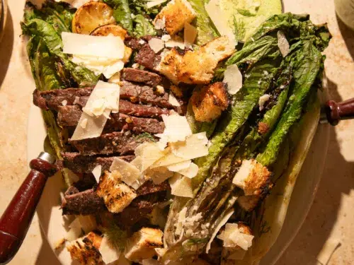 Close-up of charred romaine hearts topped with ribeye steak slices, parmesan, croutons, and a drizzle of creamy herb dressing.
