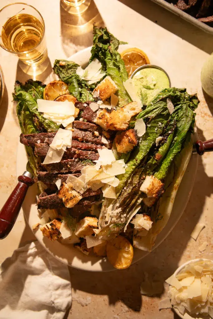 Close-up of charred romaine hearts topped with ribeye steak slices, parmesan, croutons, and a drizzle of creamy herb dressing.