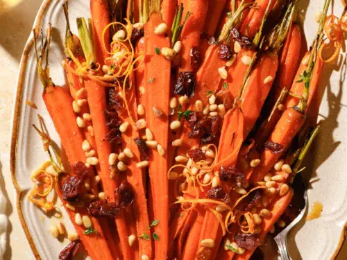 Overhead view of roasted honey glazed carrots topped with dried cranberries, toasted pine nuts, and orange zest on a serving platter.