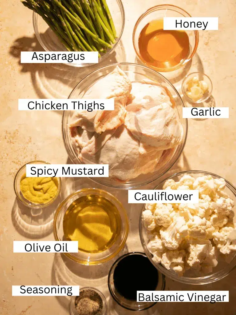 Overhead view of raw chicken thighs, asparagus, cauliflower, balsamic vinegar, honey, mustard, garlic, and seasonings laid out in bowls.
