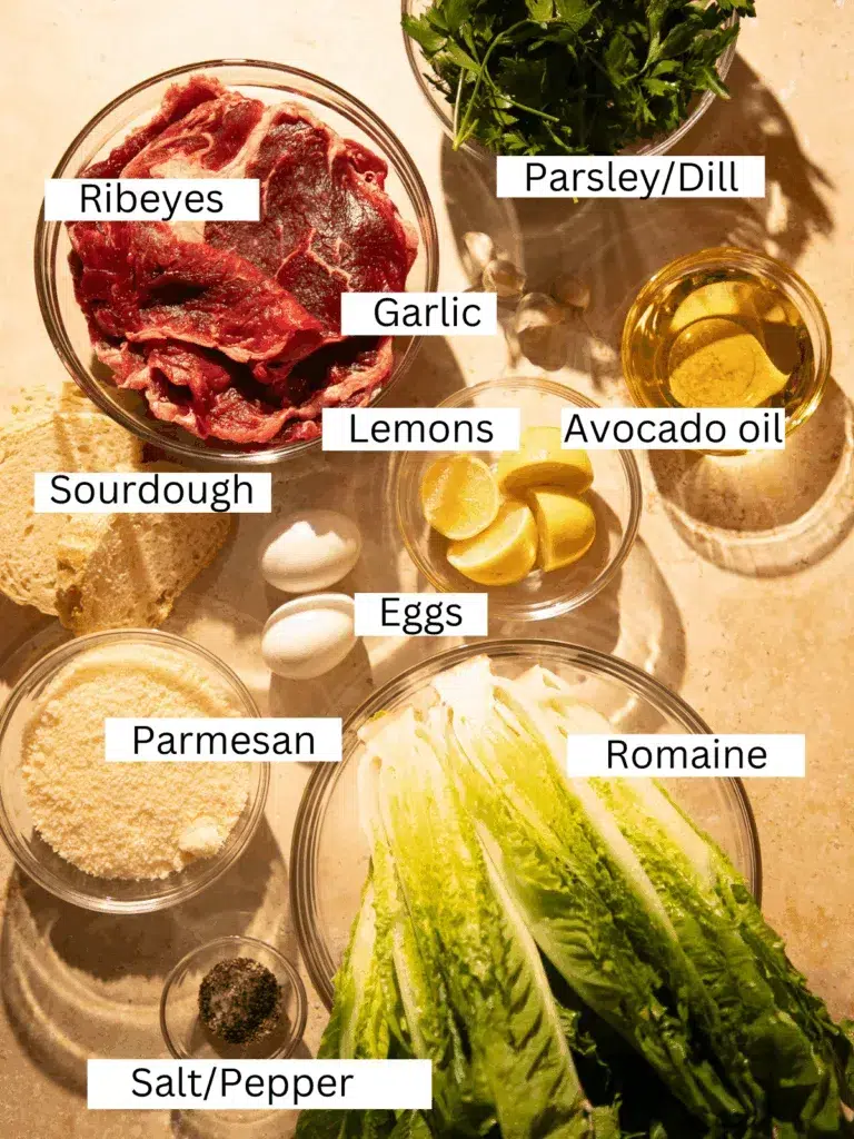 Overhead view of ingredients for grilled Caesar salad including ribeye steaks, romaine lettuce, sourdough bread, lemons, parmesan, garlic, eggs, fresh herbs, and seasonings.
