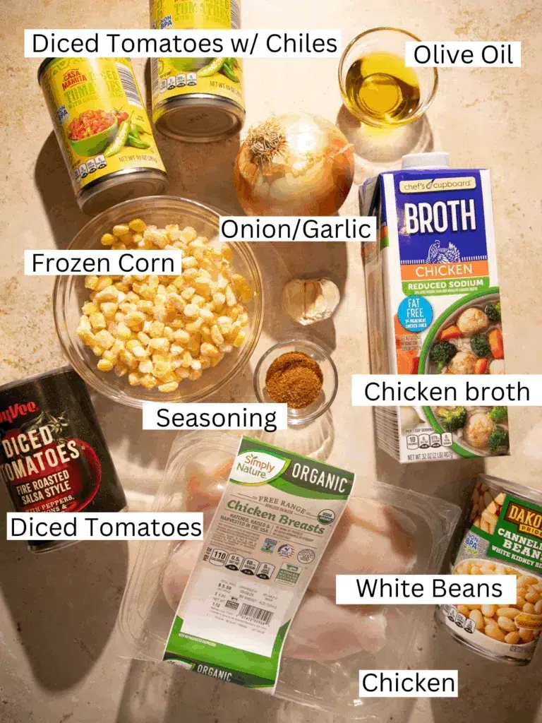 Overhead view of chili ingredients including chicken breasts, canned white beans, diced tomatoes with chiles, onion, garlic, frozen corn, chicken broth, olive oil, and spices.