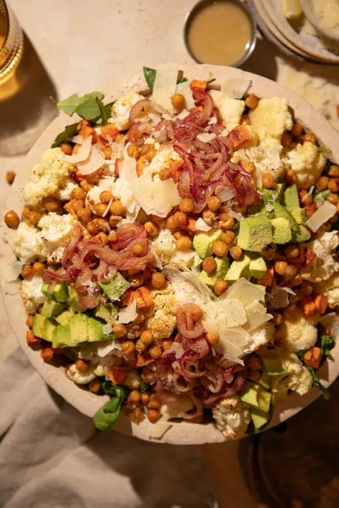 Large bowl of roasted cauliflower salad with sweet potatoes, crispy chickpeas, avocado, arugula, and parmesan with lemon tahini dressing.