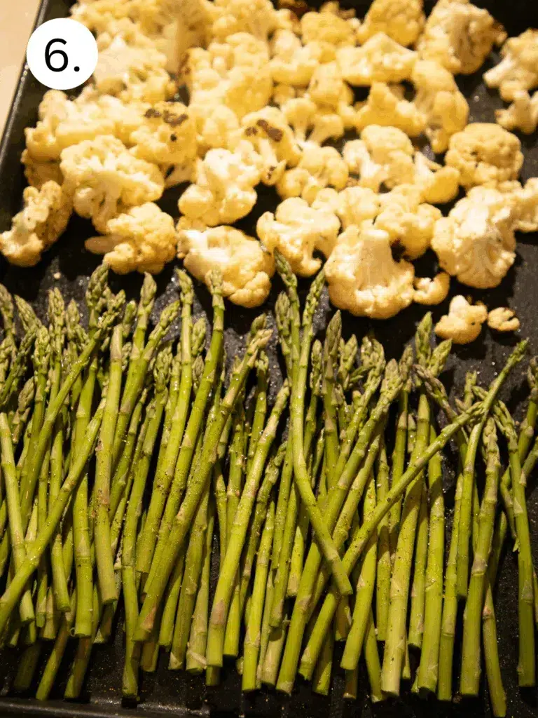 Asparagus spears arranged next to cauliflower florets on a sheet pan, ready to roast.