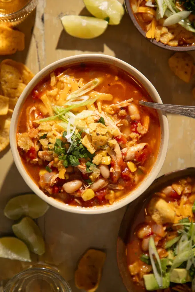 Bright bowl of chili topped with avocado chunks, shredded cheese, green onion, sour cream, and crushed tortilla chips.