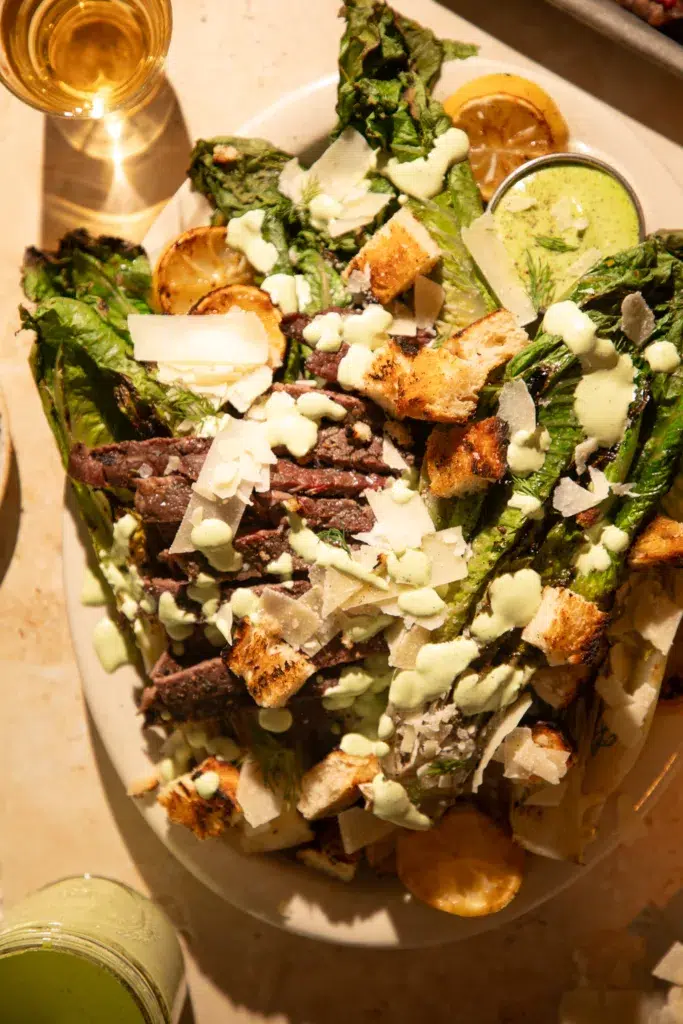 Side view of grilled Caesar salad with steak, lemon halves, and fresh parmesan shavings.