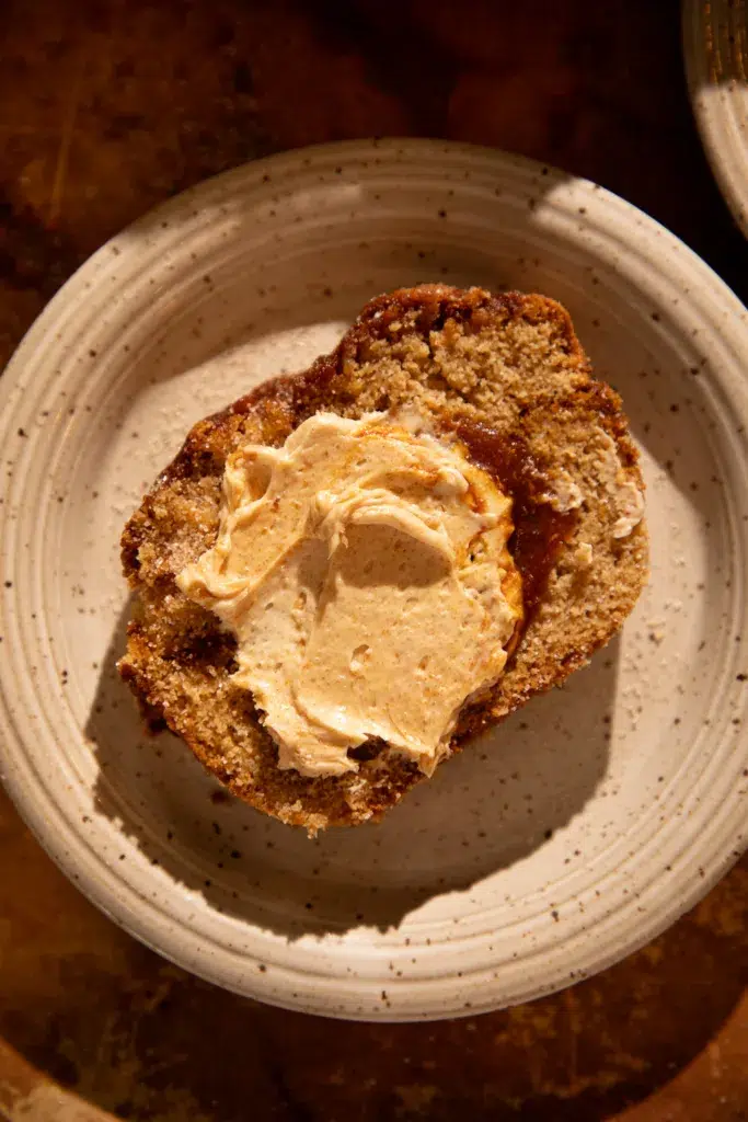 Slice of caramel apple cider donut bread topped generously with whipped maple butter on a speckled plate.