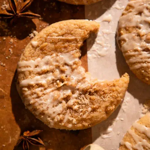 Glazed apple cider coffee cake cookie with bite missing on parchment paper with spices around.
