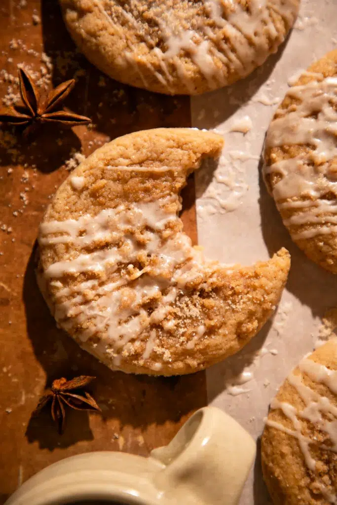Glazed apple cider coffee cake cookie with bite missing on parchment paper with spices around.