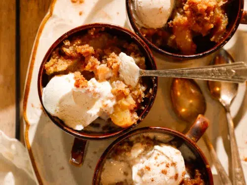 Three mugs of apple dump cake with scoops of vanilla ice cream on a rustic tray.