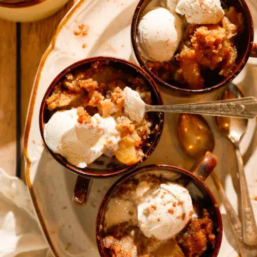 Three mugs of apple dump cake with scoops of vanilla ice cream on a rustic tray.