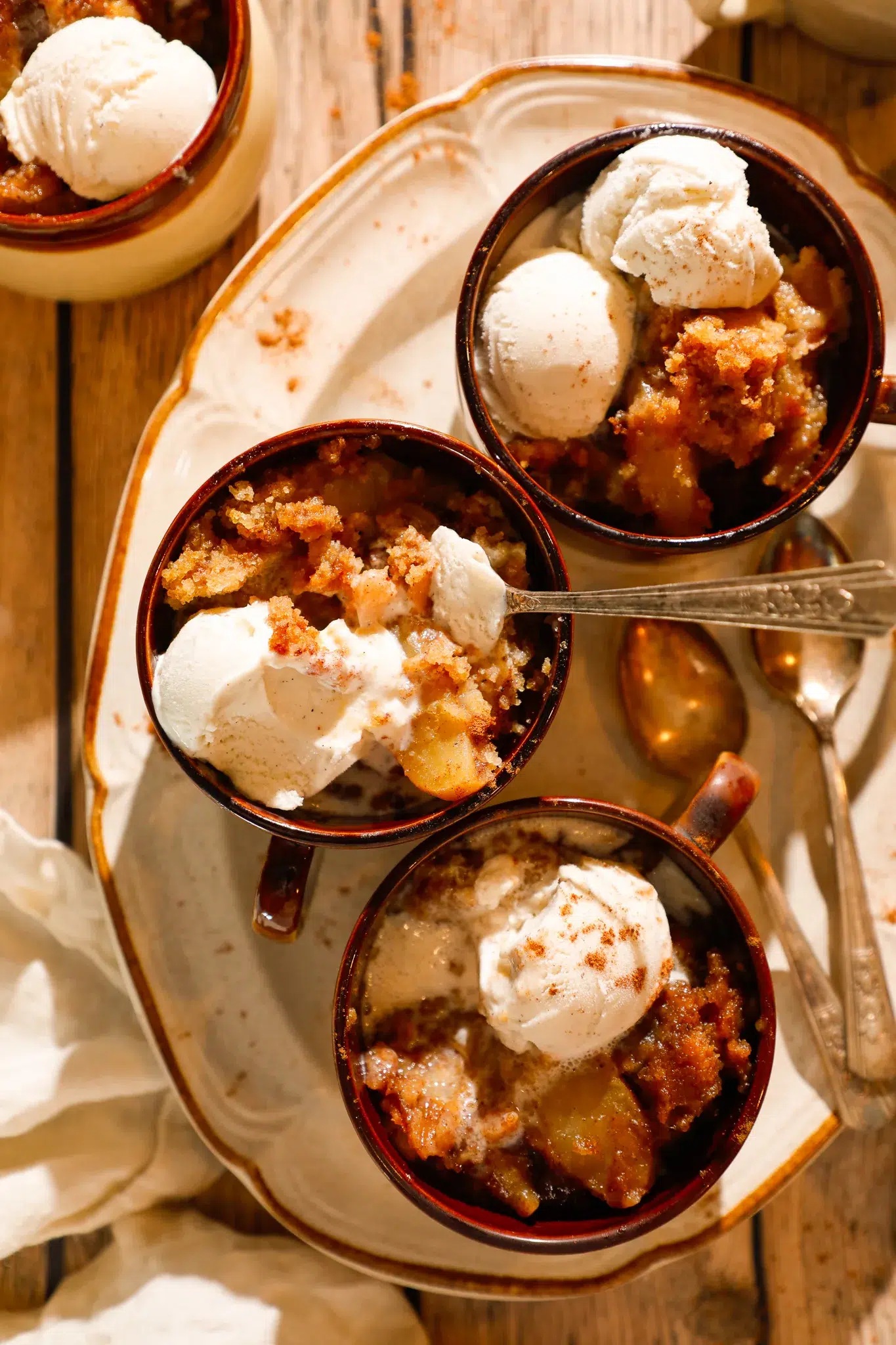 Three mugs of apple dump cake with scoops of vanilla ice cream on a rustic tray.