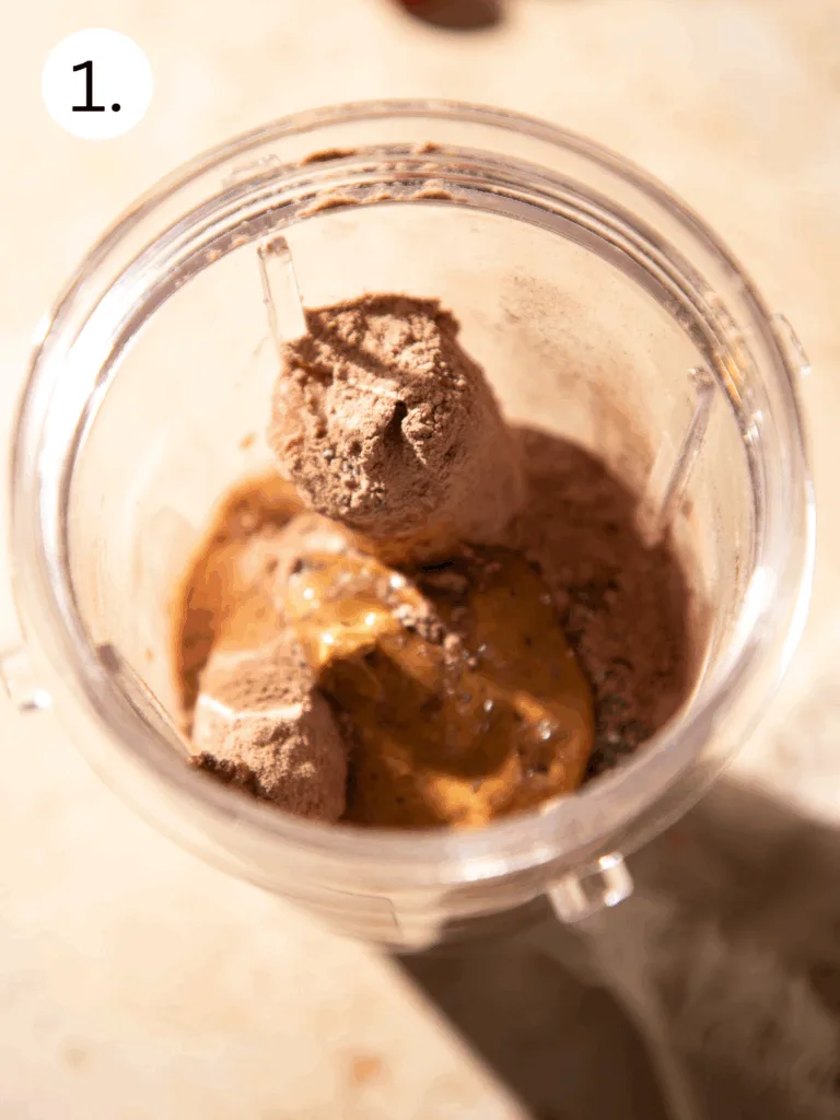 Protein powder, peanut butter, chia seeds, and other ingredients added to a blender cup to make a chocolate cherry espresso protein shake.