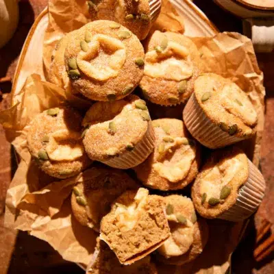Warm apple cider donut muffins arranged in a rustic pile, coated in cinnamon sugar and sprinkled with pepitas.