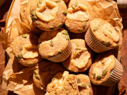 Warm apple cider donut muffins arranged in a rustic pile, coated in cinnamon sugar and sprinkled with pepitas.