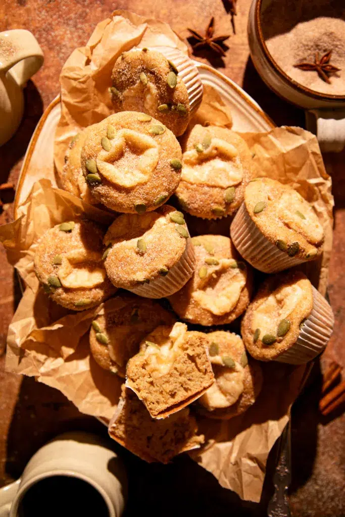 Warm apple cider donut muffins arranged in a rustic pile, coated in cinnamon sugar and sprinkled with pepitas.
