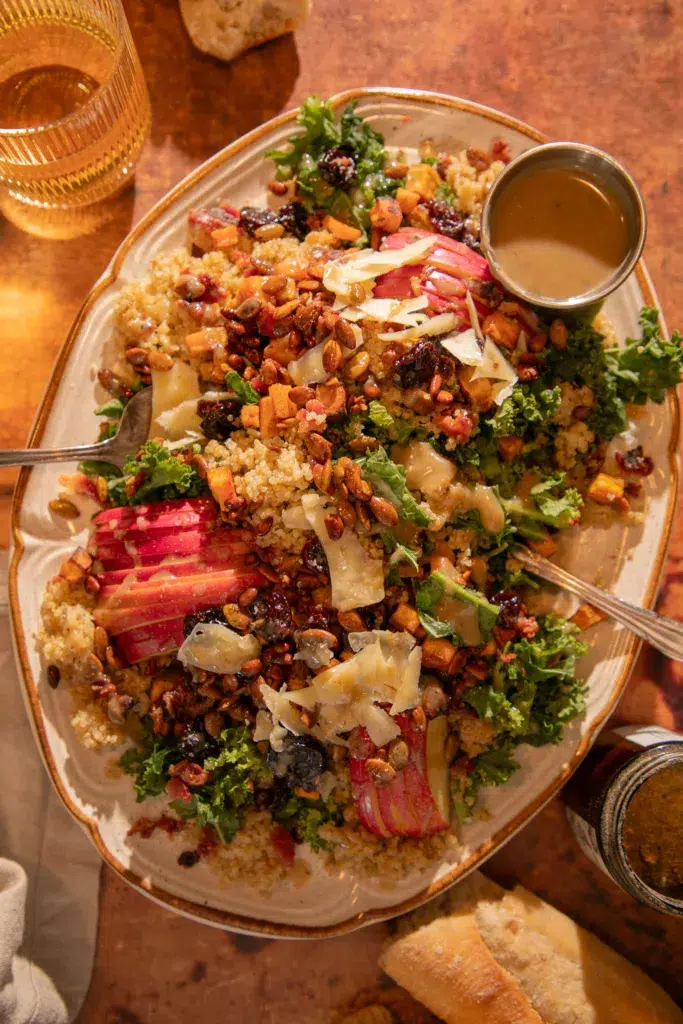 Platter of kale and quinoa harvest salad topped with roasted sweet potatoes, apples, dried cherries, nuts, and parmesan, drizzled with bacon-tahini vinaigrette.