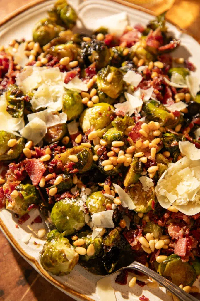 Overhead shot of roasted Brussels sprouts topped with crispy bacon, toasted pine nuts, and shaved parmesan on a serving platter.