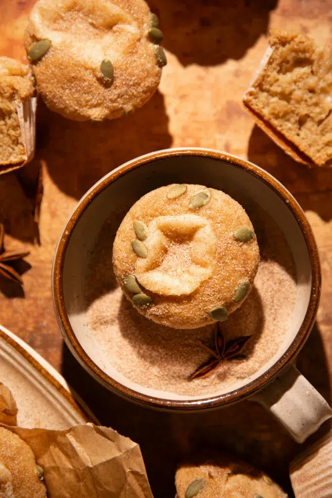 Apple cider donut muffin resting in a bowl of cinnamon sugar, topped with pepitas and a creamy filling center.