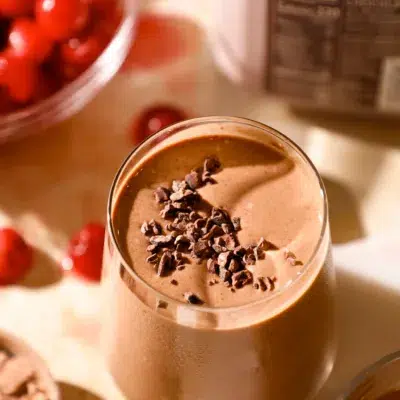 Close-up of a chocolate cherry espresso protein shake in a chilled glass, garnished with cacao nibs and surrounded by ingredients.