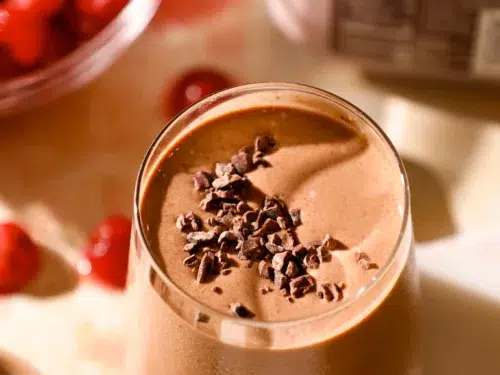 Close-up of a chocolate cherry espresso protein shake in a chilled glass, garnished with cacao nibs and surrounded by ingredients.