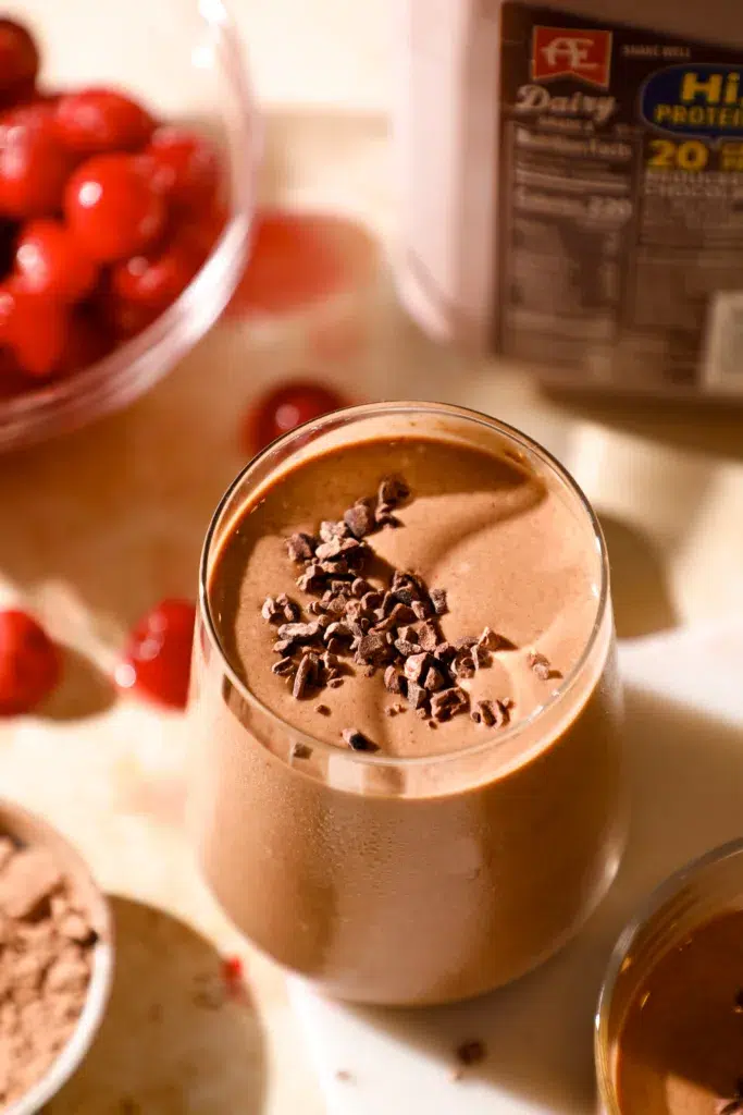 Close-up of a chocolate cherry espresso protein shake in a chilled glass, garnished with cacao nibs and surrounded by ingredients.