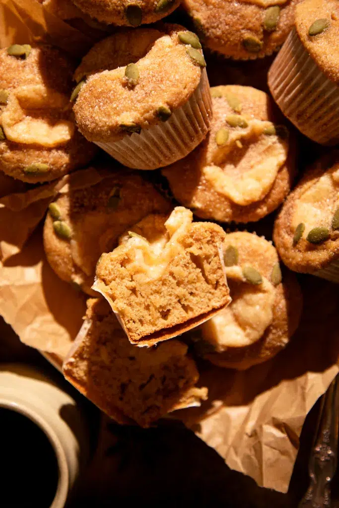 Pile of apple cider donut muffins on parchment paper with cinnamon sugar pepita topping, one muffin cut open to reveal creamy filling.