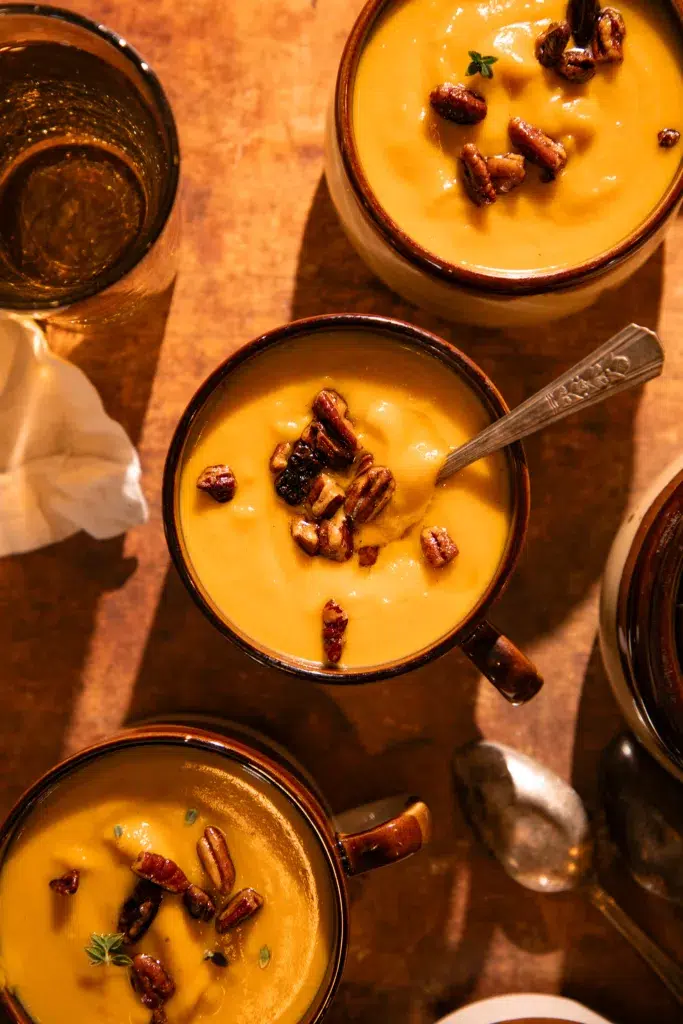 Bowls of golden butternut squash soup garnished with pecans on a wooden table.