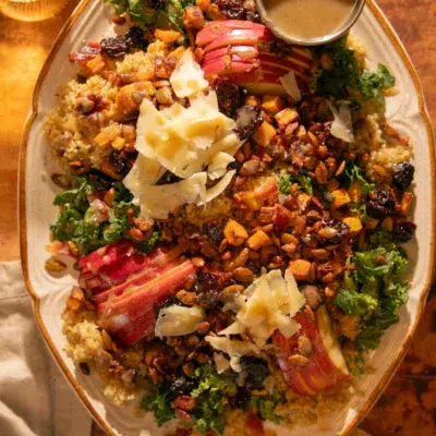Close-up of autumn harvest salad with kale, quinoa, roasted sweet potatoes, apples, dried cherries, bacon, pepitas, pine nuts, and creamy bacon-tahini dressing.