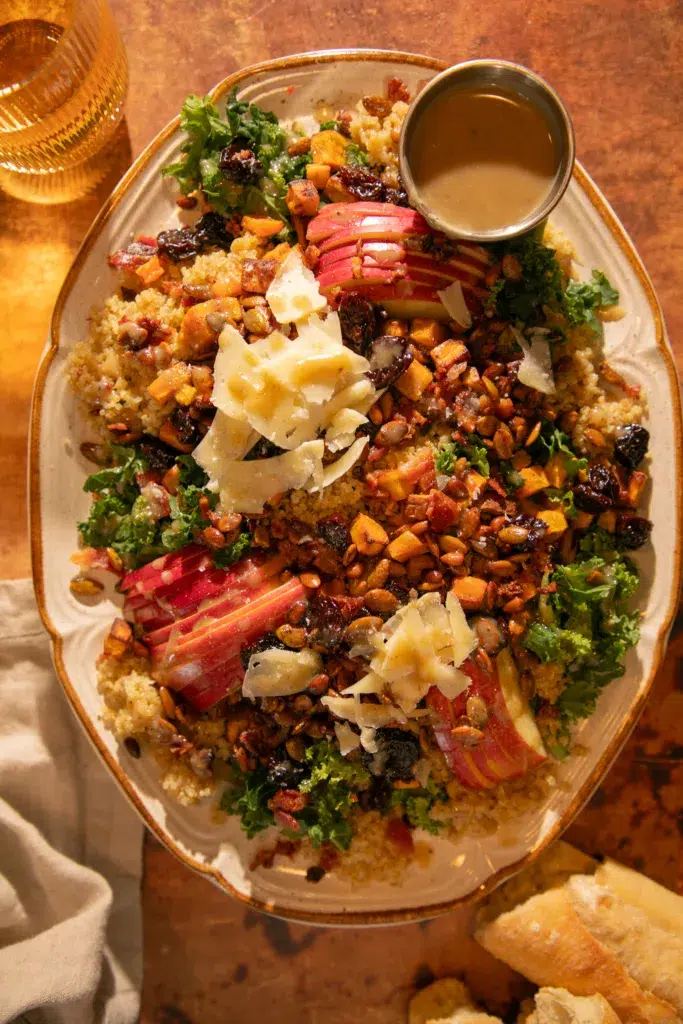 Close-up of autumn harvest salad with kale, quinoa, roasted sweet potatoes, apples, dried cherries, bacon, pepitas, pine nuts, and creamy bacon-tahini dressing.