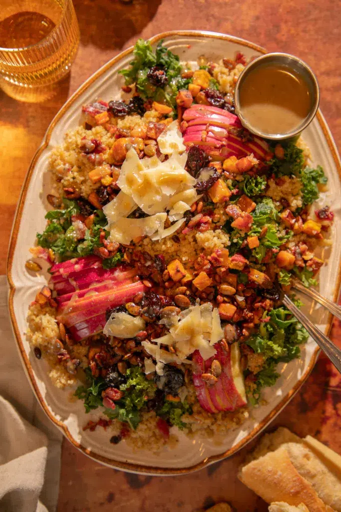 Large platter of fall harvest salad with kale, quinoa, roasted sweet potatoes, bacon, apples, dried cherries, pepitas, pine nuts, and shaved parmesan served with bacon-tahini vinaigrette.