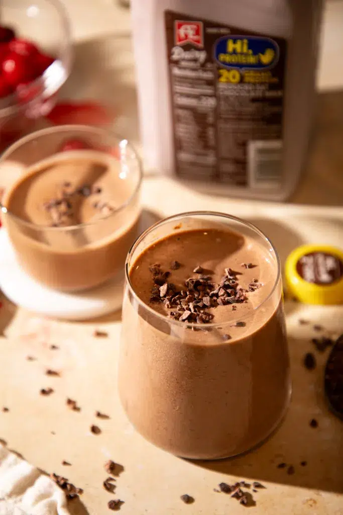Rich and creamy chocolate cherry espresso protein shake topped with cacao nibs, with cherries and chocolate milk in the background.