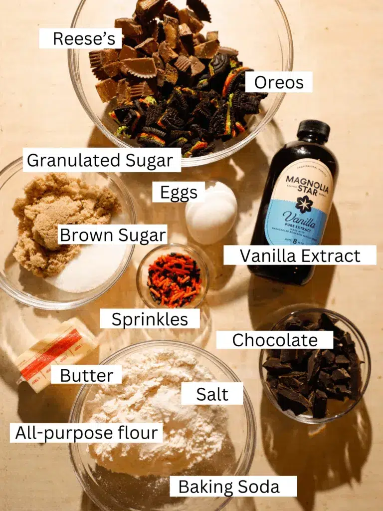 Overhead shot of ingredients for loaded Halloween chocolate chip cookies including Reese’s, Oreos, chocolate chunks, sprinkles, and baking staples.