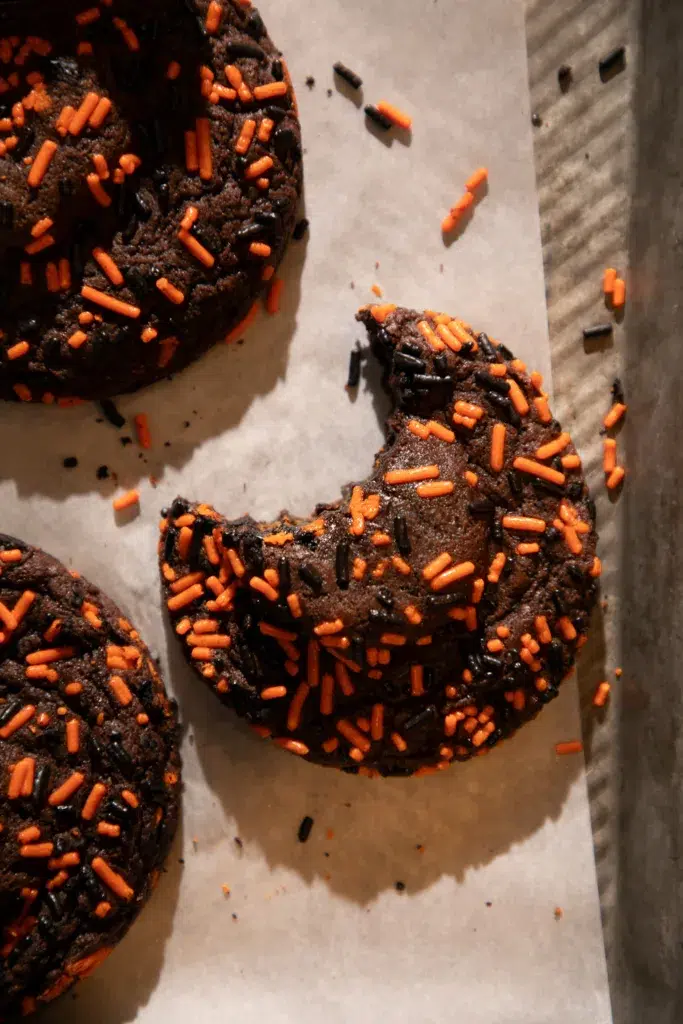 Close up of a chocolate Halloween cookie with festive sprinkles and a bite taken out to show soft inside texture.