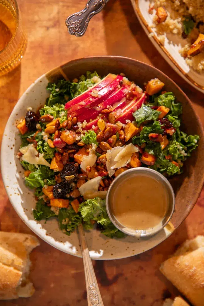 Individual serving of kale and quinoa fall harvest salad with sliced apples, roasted sweet potatoes, dried cherries, and toasted pepitas, served with a side of bacon-tahini vinaigrette.