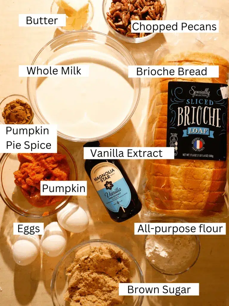 Overhead view of ingredients including brioche bread, pumpkin purée, brown sugar, eggs, whole milk, butter, chopped pecans, flour, pumpkin pie spice, and vanilla extract.