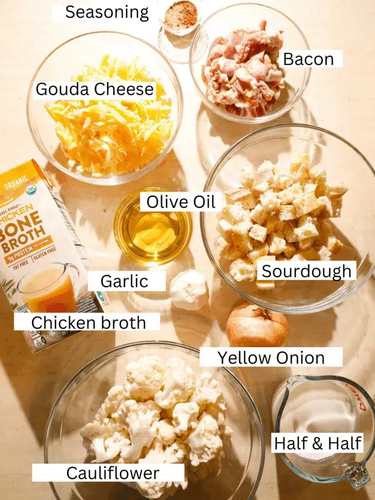 Overhead view of ingredients including cauliflower, gouda cheese, bacon, roasted garlic, onion, chicken broth, olive oil, and sourdough cubes