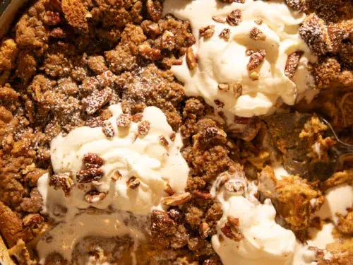 Pumpkin brioche French toast bake with pecans, partially scooped, showing soft custardy inside topped with whipped cream and streusel.